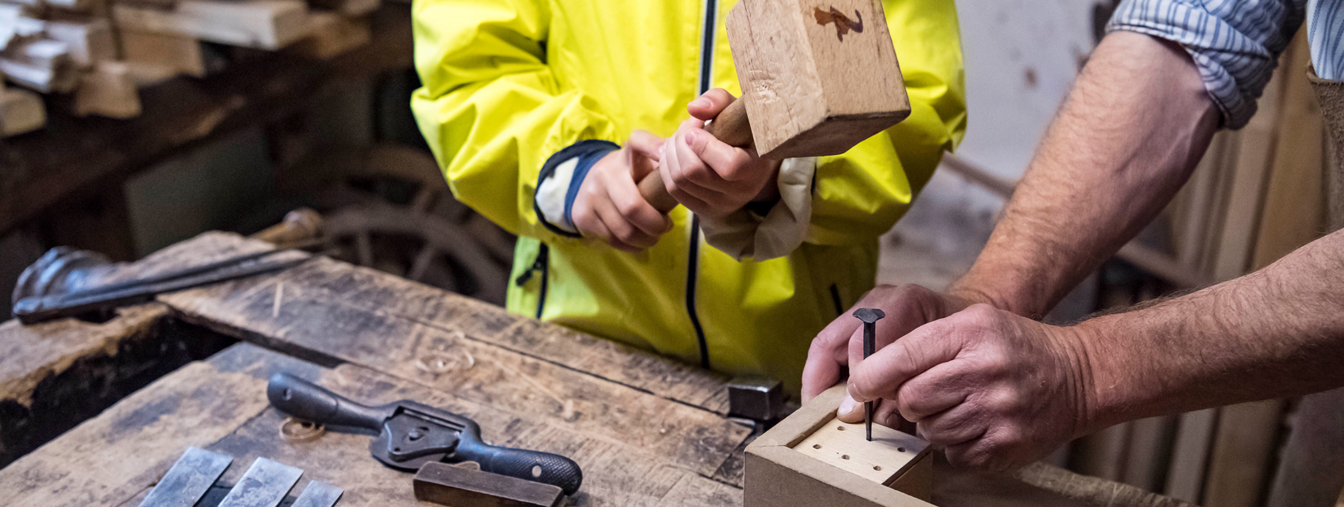 Kind schlägt mit Holzhammer auf einen Nagel, der von einem Mann auf einem Holzstück gehalten wird